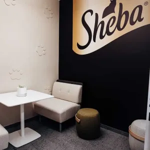 Kleiner Meetingraum mit Sheba-Logo, hellen Sitzmöbeln und Akzentwand in Schwarz-Gold